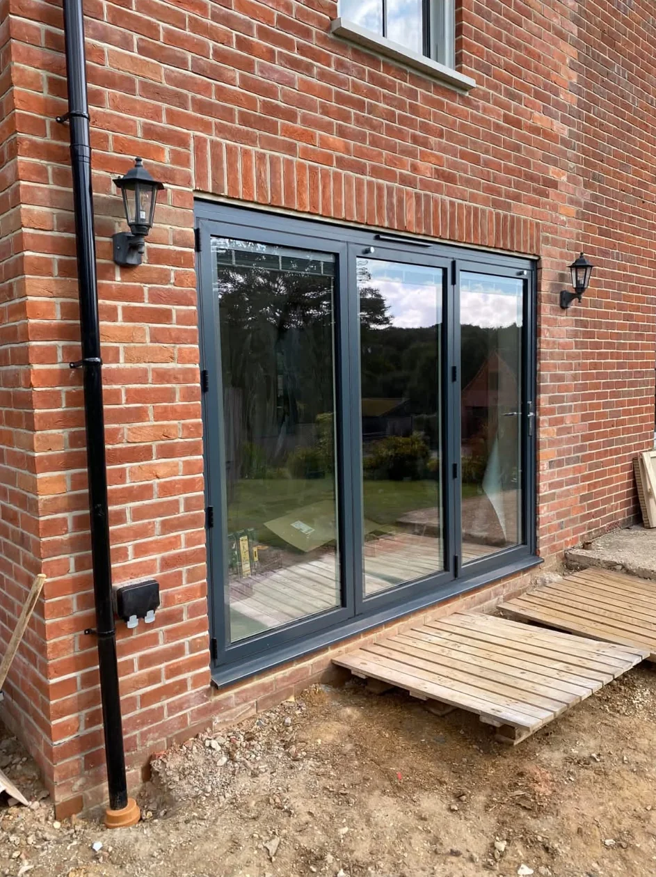 A dark grey three-pane bifold door in a red-brick rear wall, between two wall lanterns
